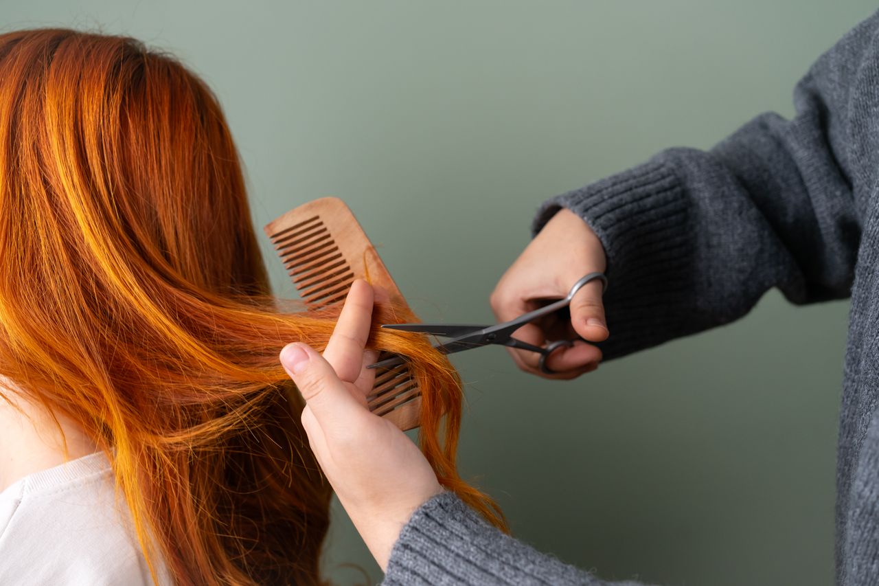 Close-up of red hair being trimmed with scissors and comb on green background.
