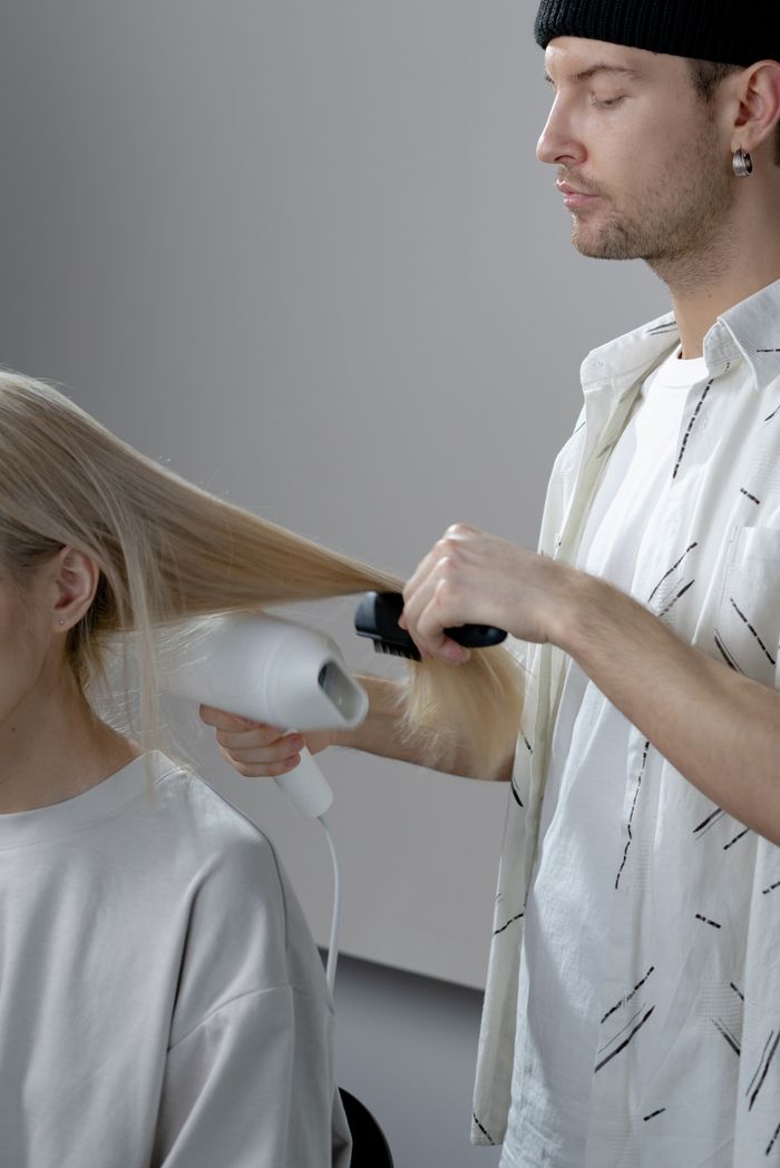contact-img Hairstylist uses blow dryer and brush to style client's long blonde hair in salon.